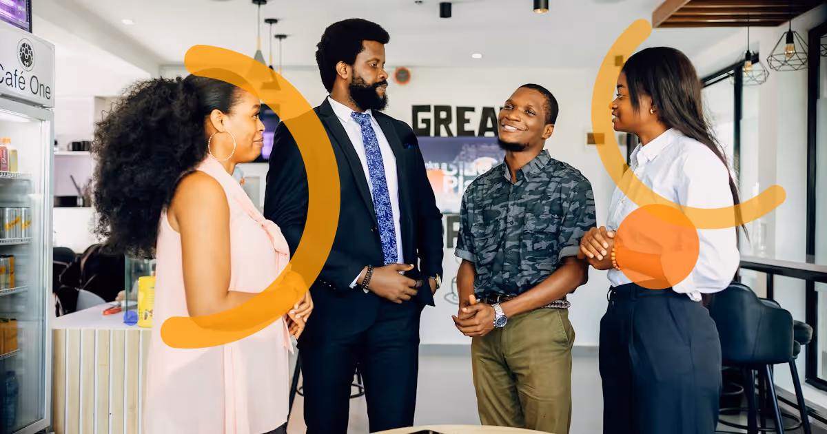 Four colleagues standing around a small table in a bright office café, smiling as they welcome a new team member.